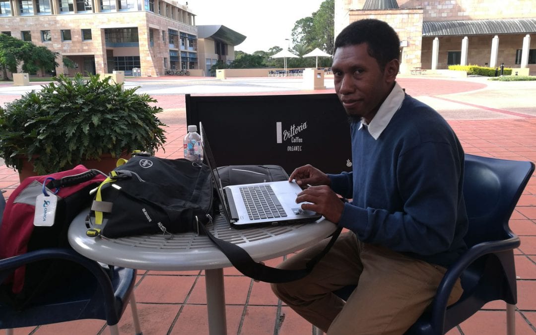 Emmanuel Powuh David is seated with his laptop at a table between buildings at Bond University